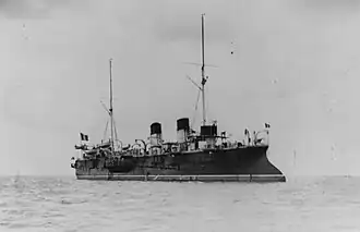 A large, black ship with two funnels and two thin masts sits at anchor in a calm sea.