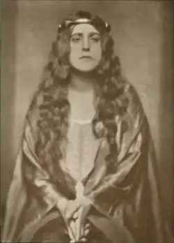 A white woman with long light hair, wearing a loose robe and a circlet, hands clasped low in front of her