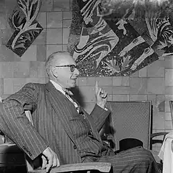 George Szell, seated, in a pin-stripe suit, gesturing with his left hand