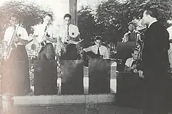 From left to right: three standing saxophone players, one sitting guitar player, one accordion player, one fiddler, one conductor and one barely visible drummer. All are men and wear six-pointed stars.