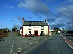 Front elevation of a white-painted two-storey rural pub with a central door, two lower and two upper windows edged with red, situated between two roads under a mostly-blue sky