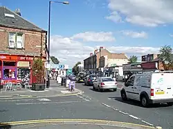 The junction of High Street and Church Road. A terrace of stone fronted buildings, with a curved corner building fronted in white, from the mid-late 19th century. The modern shop fronts are not in keeping the original style of the buildings. It is set back from the road by modern paving. The far section of the building is the old fire station building (formerly Urban District offices) are .