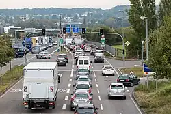 Vehicles backed up on a divided roadway seen from above. In the foreground is a traffic signal; there are blue signs in German further down the road as it narrows. In the rear is a developed hillside, partially obscured by bluish haze