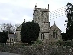 Grey stone building on 3 bays with a square stone tower at near end of central bay. To the left is a porch with slate roof. In front is a yew tree and gravestones behind a stone wall separating it from a road.