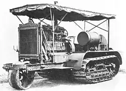 An early bulldozer-like tractor, on crawler tracks, with a leading single wheel for steering - projecting from the front - on an extension to the frame. The large internal combustion engine is in full view, with the cooling radiator prominent at the front. An overall roof is supported by thin rods, and side protection sheeting is rolled up under the edge of the roof.