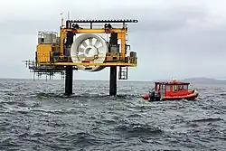 Photo taken at sea of the OpenHydro turbine, raised above the water supported between 2 metal piles. There is a walkway around the turbine for maintenance access and a small orange boat nearby. The turbine has an outer and inner ring, with 10 blades between.
