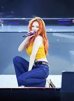 A red haired woman wearing a yellow top and blue jeans performing on stage while holding a microphone in her right hand.