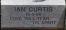 A grayish stone block with "Ian Curtis 18-5-1980 Love Will Tear Us Apart" carved into it in a sans-serif typeface