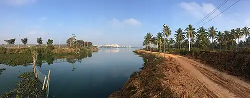 A water body surrounded by a mud path and trees. In the far distance apartments are visible.