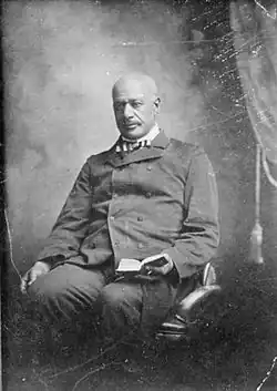 An older, balding black man with mustache, wearing a double-breasted suit and striped bow tie. He is sitting in a chair holding an open book in his left hand.