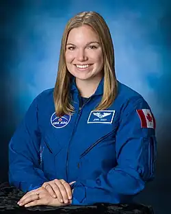 Gibbons, wearing her blue Canadian Space Agency uniform bearing the flag of Canada, smiles at the camera, her hands folded.