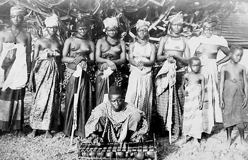 Photographie d'un groupe de femmes partiellement dénudées, fixant l'objectif. Un homme assis au premier plan joue d'un instrument de musique.