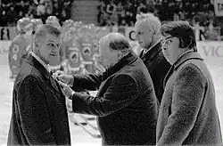 Black and white photo of four men wearing suits while standing on the ice of a hockey rink during am awards ceremony