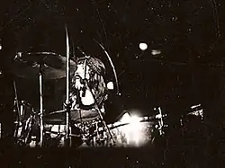 A black and white photograph of John Bonham wearing a headband and behind the cymbals of a drum kit