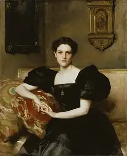 A seated woman wears a dark dress with puffed sleeves. Her hands are clasped in her lap, and she gazes directly at the viewer. She sits on a sofa with a patterned cushion. Behind her, a wall with two framed artworks or decorative pieces.