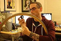 A man with glasses is removing the spokes from a bicycle wheel in an office.