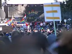 Photo of the stage and the crowd. Karácsony is giving his speech on the stage, and people in the crowd hold Pride flags and a sign reading "Proud asexual / Shameful Hungarian".