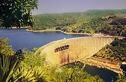An impressive dam, built in a semi-circular shape, viewed from a vantage point above and to the right of its front. A large lake can be seen on the other side of the dam as well as a river before it.
