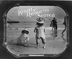 Photo shows a framed paper print of a funny motion picture lantern slide depicting children at a beach overlooking the ocean, one of whom is standing, with the caption, "Please remain seated."