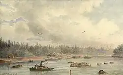 a watercolour painting of a small steamboat towing two barges piled high with equipment and three large canoes filled with men. Other barges and canoes are visible near the shore behind. A single building is visible near the left-hand side of the frame. The land is densely forested. There are small groups of men and canoes busy on shore and further along the river. Gulls wheel overhead against a sky where white clouds drift by.