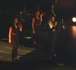 Three women sing on stage. They wear similar outfit (a dark blouse and trousers).