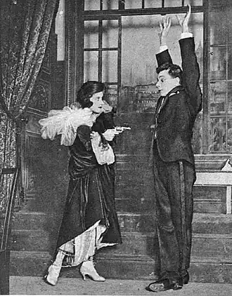 Stage scene with young white man, hands in air, being threatened by a young woman with a pistol