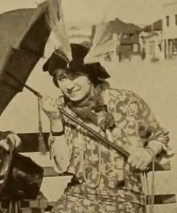 A young white woman seated outdoors, holding an umbrella and wearing a floral-print dress and a hat with plumes