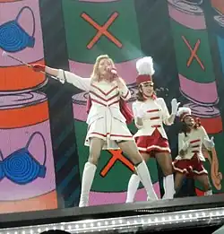 A blond woman standing in front of a black and red wall, wearing a black dress, and with her hands on her waist.