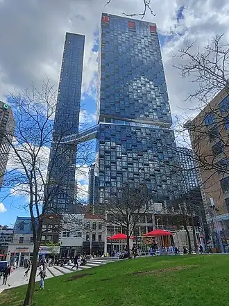 Two large towers in different stages of construction, connected by a sky bridge. Pedestrians mill about on a plaza in the foreground.