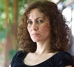 Head shot of Maha Haj, she has shoulder-length curly brown hair. The background is blurry trees