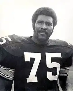 A man with an Afro and a beard in a black football uniform with 75 written in white