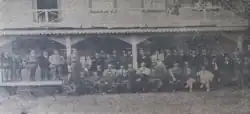 A black-and-white photograph of a group of men in early 20th century formal attire standing in front of a building