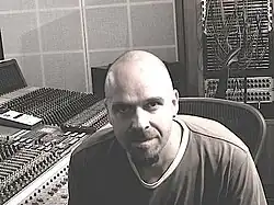 In a de-saturated recording studio, a bald, white male glares at the viewer with a faint smile