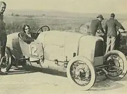 Black and white photo of a white woman in an old race car. Several men stand around.