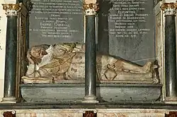 This image shows the elaborate 17th-century monument to Sir Lionel Tollemache in St Mary’s Church, Helmingham, Suffolk. The monument features a life-sized, painted recumbent effigy of Sir Lionel lying on his side, propped up on his left elbow in a relaxed, almost conversational pose. He wears ornate plate armour with gold detailing, a lace ruff at his neck, and a sash across his chest. His helmet rests beside him, and his facial features — neatly trimmed beard and moustache, long wavy hair, and a calm expression — are carefully painted. The armour is decorated with gilded accents, including rosettes and straps. The effigy lies beneath an architectural canopy framed by black columns with gold Corinthian capitals. Behind him is an inscribed black tablet with Latin text commemorating his life and service. The text records his loyalty to King James I and King Charles I, his role as Vice-Admiral of Suffolk, and his death in 1640 at the age of 49. The style, with its reclining pose, rich gilding, and lifelike painting, reflects the grand funerary traditions of English nobility in the early Stuart period.