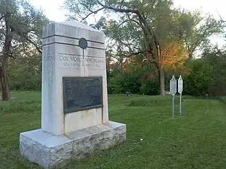 An angled view of a large stone monument with a placard and two nearby white historical markers.