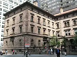The north wing of the Villard Houses as seen from across Madison Avenue. The facade is made of brown stone and has rectangular window openings. To the right is a black fence, behind which is the courtyard.