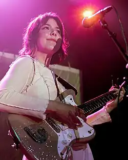 Nanna Bryndís Hilmarsdóttir wearing a white long-sleeved top with a white vest, smiling and playing electric guitar in front of a microphone onstage