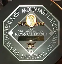 A black circle with an octagonal read "KENESAW MOUNTAIN LANDIS MEMORIAL BASEBALL AWARD". In the middle of the octagon is a baseball diamond which contains, from the top, Judge Landis' face in gold, "Most Valuable Player", the winner's league, his name in a gold rectangle, and his team.