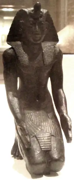 A small kneeling bronze statuette, likely Necho II, now residing in the Brooklyn Museum
