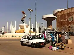 A taxi at a roundabout with an abstract sculpture. There are water towers in the background.