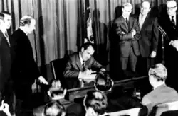 Photograph showing President Nixon seated at a desk signing the bill, with multiple people watching him; those to his right and left are standing, and those facing him in the foreground are seated