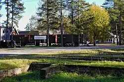 Stationary train carriages sit at the entrance of Norrbotten Railway Museum surrounded by trees on a clear, sunny day. A road and some greenery is visible in the foreground.