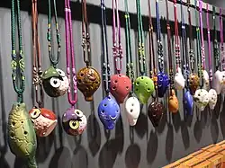 Colourful four-hole English pendant ocarinas hanging from a rail by elaborately-knotted 2- and 3-colour lanyards. Three are shaped like squat owls; several have low-relief or painted patterns.