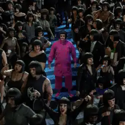 Oliver Tree as his character Cornelius Cummings, with a bob haircut and all-pink outfit, standing on a street crossing surrounded by people all with the same bob haircut