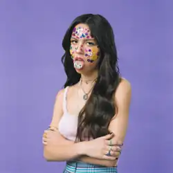 Olivia Rodrigo crossing her arms and sticking her tongue out. Her face is covered in stickers, with the ones on her tongue spelling out "SOUR".