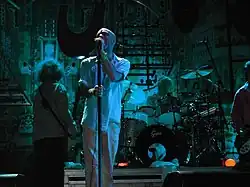 A blue-tinted photograph of musicians in front of an industrial background. From left to right: a long-haired male stands with his back to the camera playing bass guitar, a middle-aged Caucasian male sings into a microphone, a middle-aged Caucasian male plays behind a black-and-silver drum set on a riser, and a guitar player is mostly cropped from the extreme right of the photo.