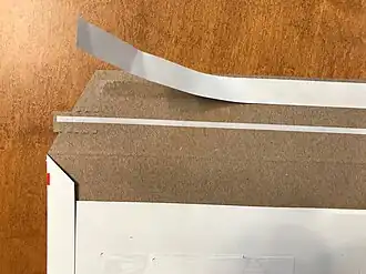 Paperboard mailing envelope showing PSA adhesive with release liner and with tear tape