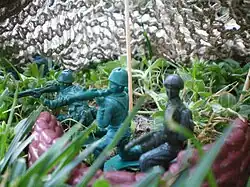 Three green plastic bazooka men on dirt in between several strands of grass