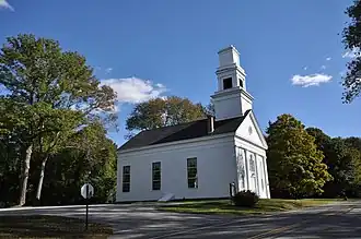Church building featuring a tall, white steeple with a pointed spire, surrounded by trees and a well-maintained lawn.